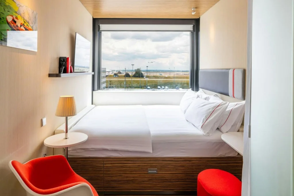 Modern compact hotel room with a large double bed, panoramic airport view, wooden furnishings and red accent chairs at citizenM Paris Charles de Gaulle