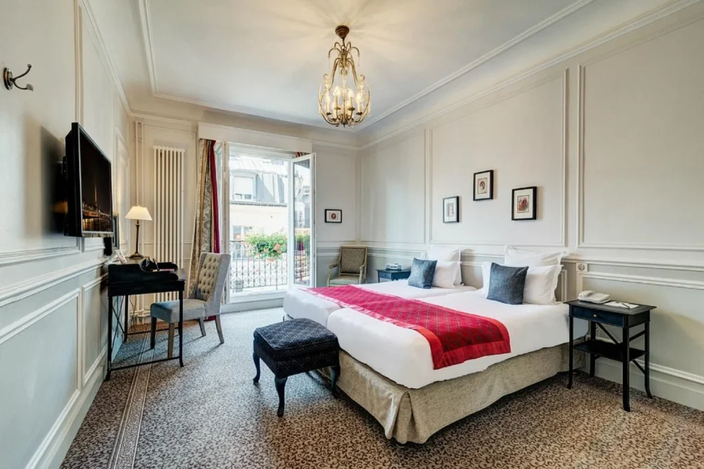 Spacious and elegant Parisian hotel room featuring a comfortable double bed, classic wall paneling, decorative chandelier, vintage-style furnishings and open French balcony doors at Hôtel Bradford Elysées - Astotel.