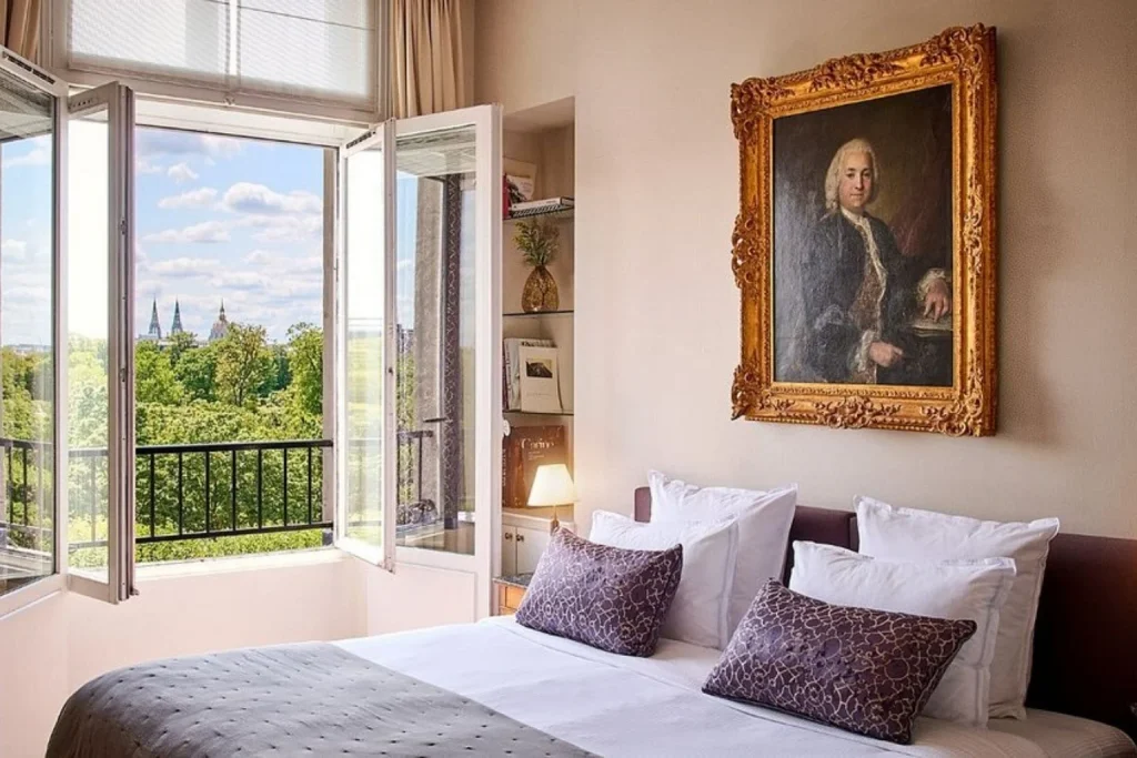 Elegant bedroom with classic décor, a large framed portrait and open windows overlooking the rooftops and greenery of central Paris at Hotel Brighton Paris.