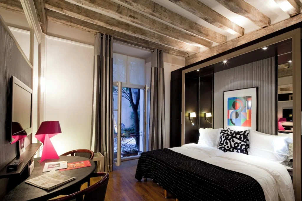 Elegant hotel bedroom with exposed wooden ceiling beams, white bedding, black accents, pink table lamp and courtyard view at Hôtel du Jeu de Paume