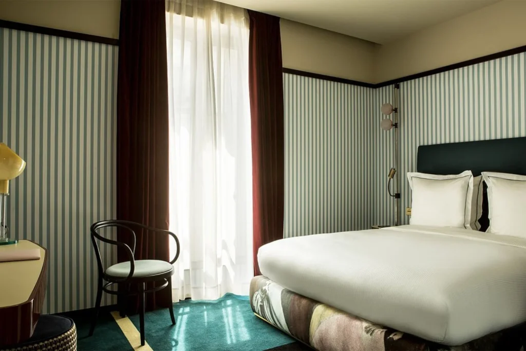 Stylish hotel room featuring blue and white striped walls, a comfortable double bed, rich teal carpeting, burgundy curtains and a classic wooden chair at Hotel du Rond-Point des Champs-Elysées.