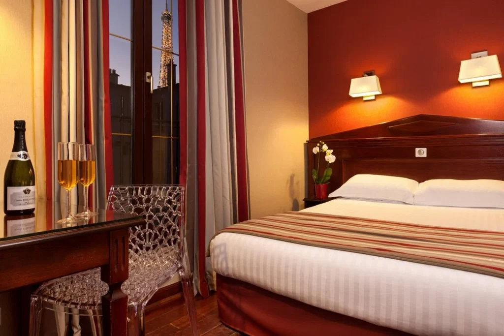 Warmly lit bedroom with red accents, champagne and a nighttime view of the illuminated Eiffel Tower at Hôtel Eiffel Rive Gauche Paris.