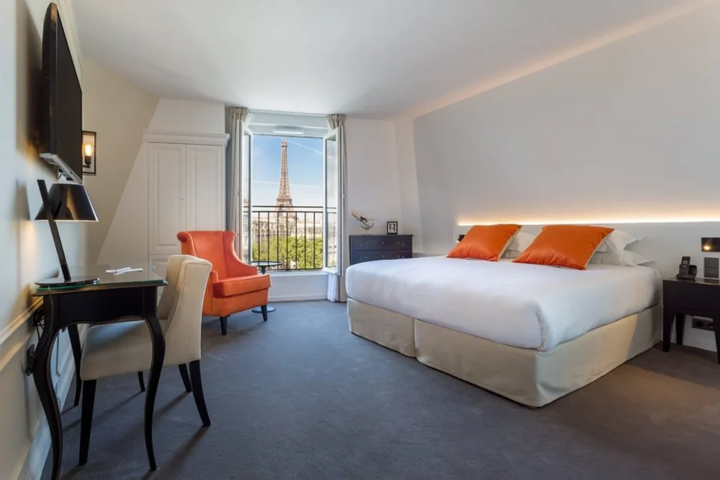 Bright contemporary bedroom with orange accents and an open window offering a direct view of the Eiffel Tower at Hôtel La Comtesse Paris.