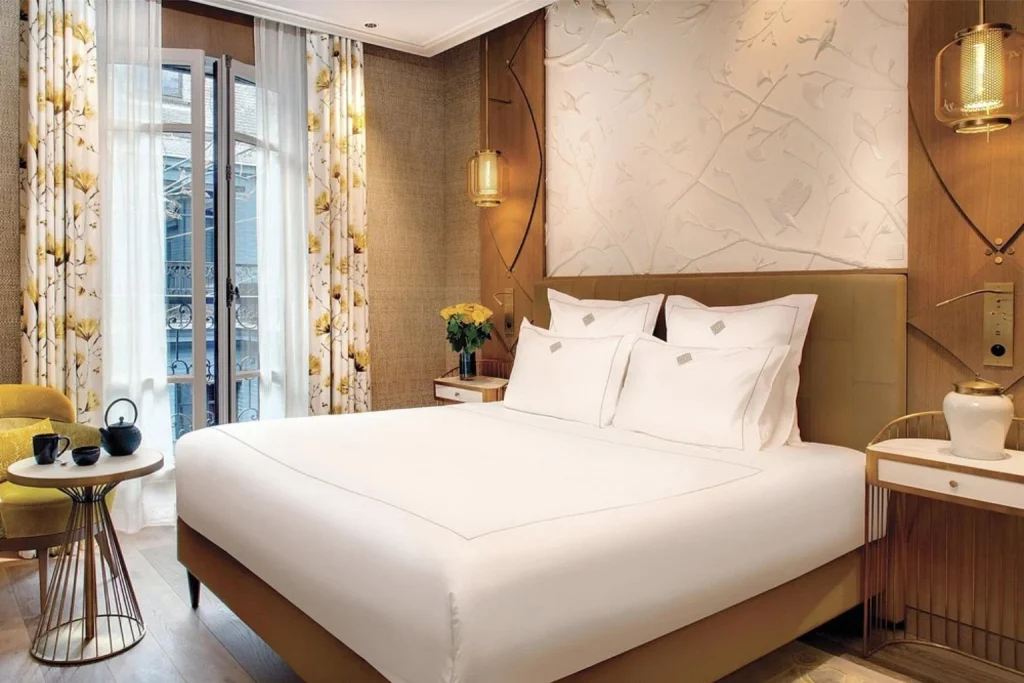 Beautifully designed Parisian hotel bedroom featuring a comfortable double bed, delicate floral curtains, soft neutral tones, elegant pendant lights and a cozy seating area at Hôtel La Demeure.