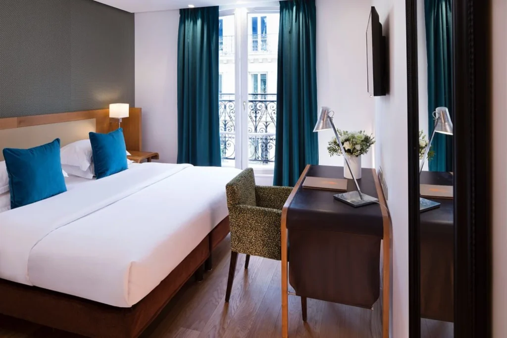 Bright and contemporary Parisian hotel bedroom featuring a comfortable double bed, vibrant blue accents, elegant wooden furnishings and tall windows overlooking a traditional Parisian building at Hotel Le Six.