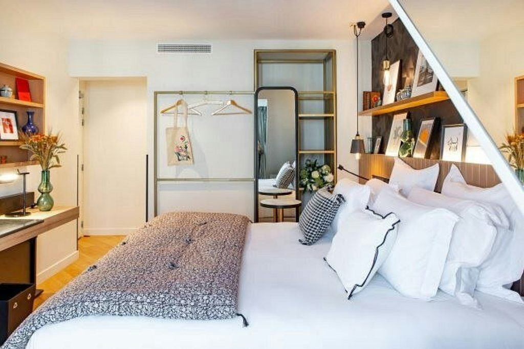 Bright and inviting boutique hotel bedroom featuring a comfortable double bed, layered cushions, contemporary wooden shelving, a full-length mirror and thoughtful decorative details at Hôtel Maison Mère.