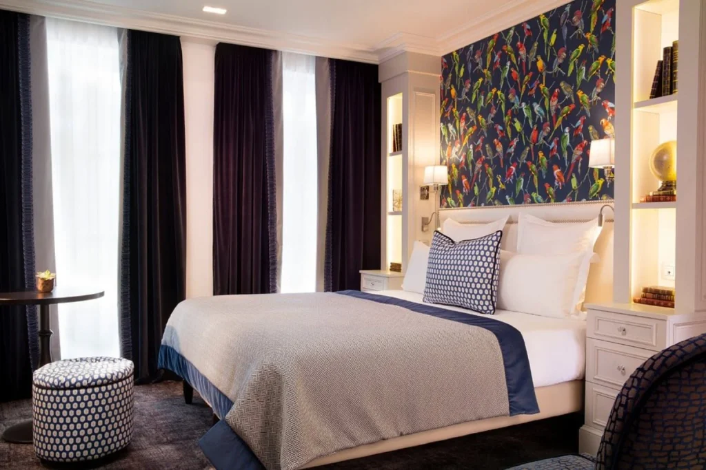 Beautifully decorated boutique hotel bedroom featuring vibrant bird-themed wallpaper, luxurious white bedding, deep navy curtains, built-in shelving and elegant decorative accents at Hotel Monge Paris.
