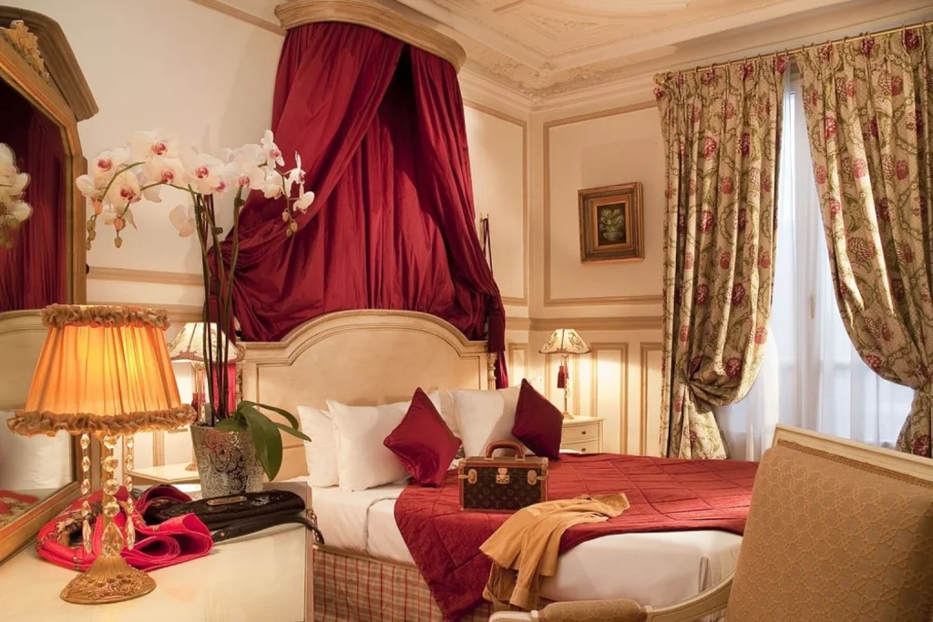 Charming classic French bedroom featuring a luxurious burgundy canopy bed, ornate floral curtains, antique-style furnishings and delicate orchid arrangements at Hôtel Résidence Henri IV.