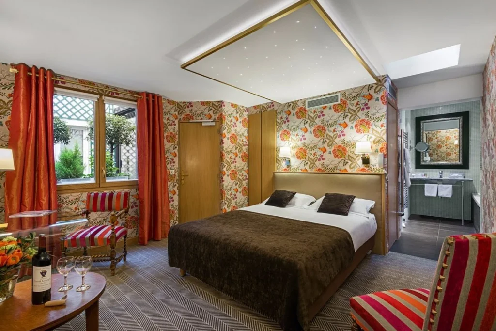 Colorful bedroom with floral wallpaper, striped armchairs and an open ensuite bathroom at Hôtel Saint-Paul Rive Gauche Paris.