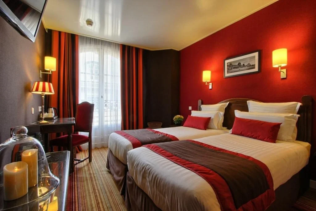 Cozy Parisian hotel room featuring two single beds, rich red walls, striped curtains, traditional wooden furniture and warm bedside lighting at Hôtel Trianon Rive Gauche.
