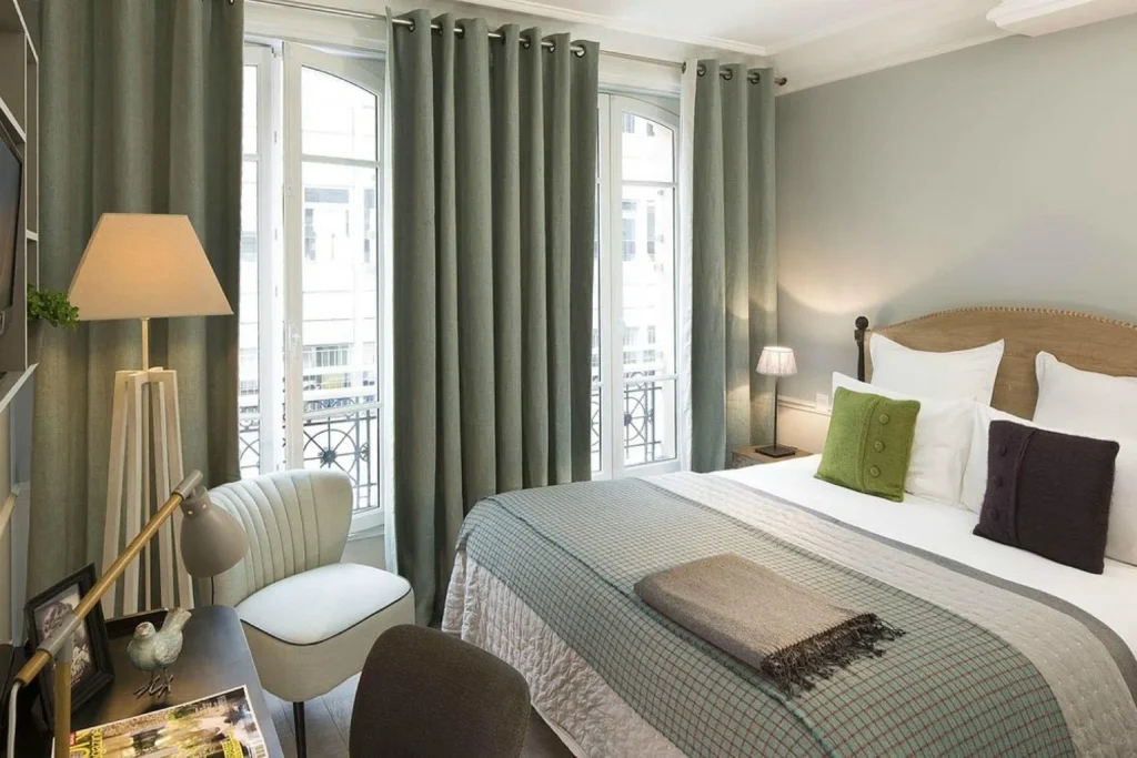 Elegant bedroom with large Parisian windows, soft green décor and a comfortable seating area at Maison Chomel Paris.
