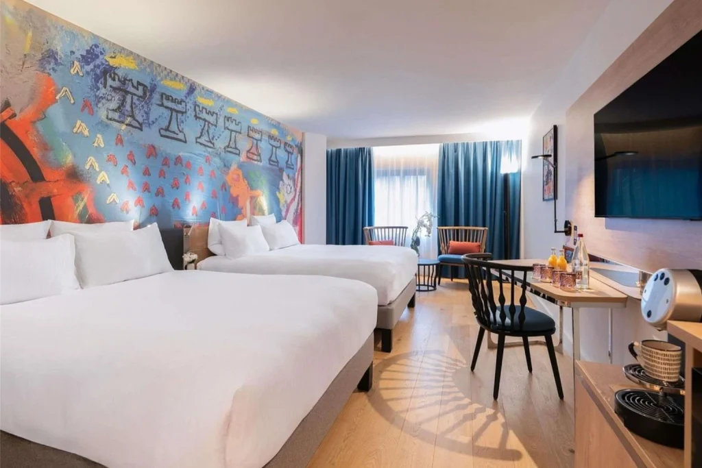 Bright and contemporary family room featuring two comfortable double beds, vibrant artistic wall decor, a seating area, wooden flooring and modern amenities at Novotel Paris Les Halles.