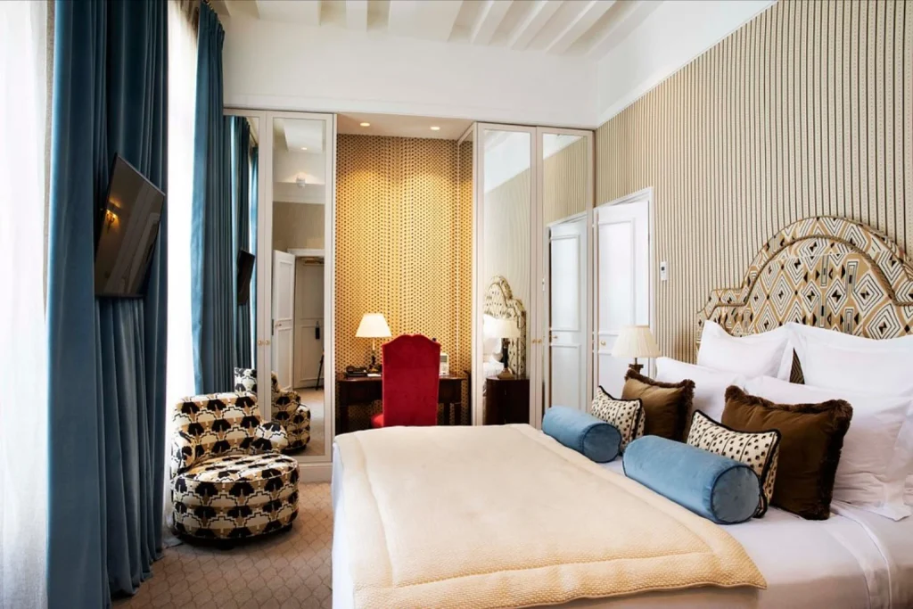 Beautifully designed luxury bedroom featuring an ornate headboard, plush bedding, blue velvet curtains and sophisticated Parisian decor at Relais Christine Paris.